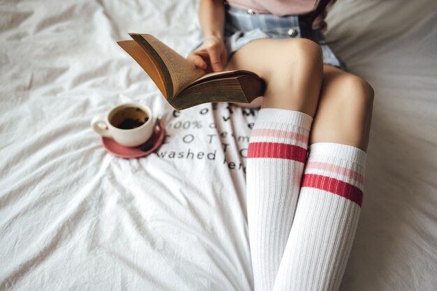 Woman lying in a white bed, drinking tea at home and reading book.