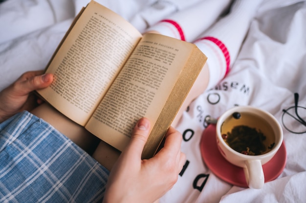 Woman lying in a white bed, drinking tea at home and reading book.