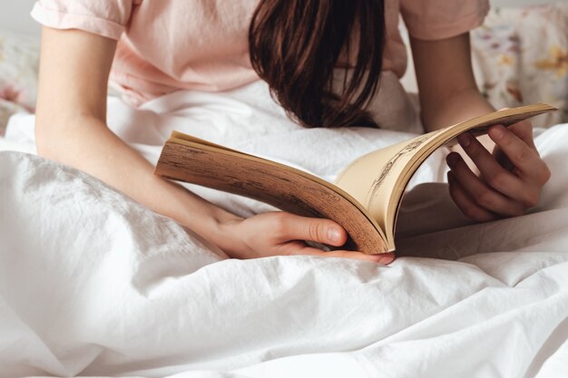 Woman lying in a white bed at home and reading book.