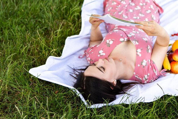 하얀 침대 시트에 누워 있는 여성은 책 노트를 잡고 읽습니다. 레이디는 도시 공원에서 휴식을 취합니다.