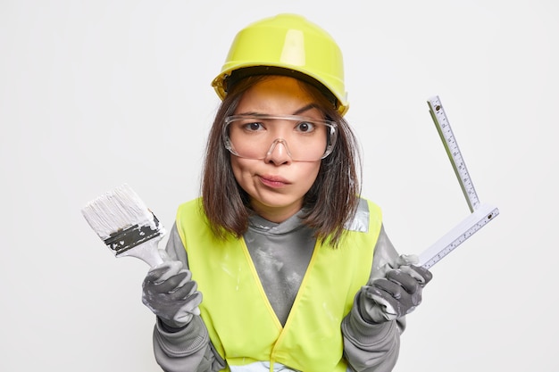 woman maintenance worker holds painting brush and tape measure smirks face wears protective hardhat and uniform isolated on white 