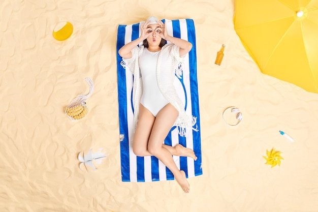 woman makes binoculars with hands on eyes wears swimhat and white bikini gets suntan at beach poses on blue striped towel surrounded by different items