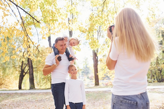 Woman makes a photo of her husband with kids in autumn park.