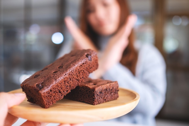 A woman making arms cross sign to refuse a brownie cake in wooden plate from someone