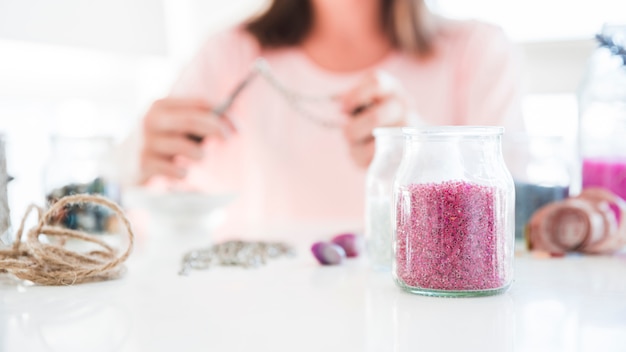 A woman making handmade jewelry and pink beads jar on desk