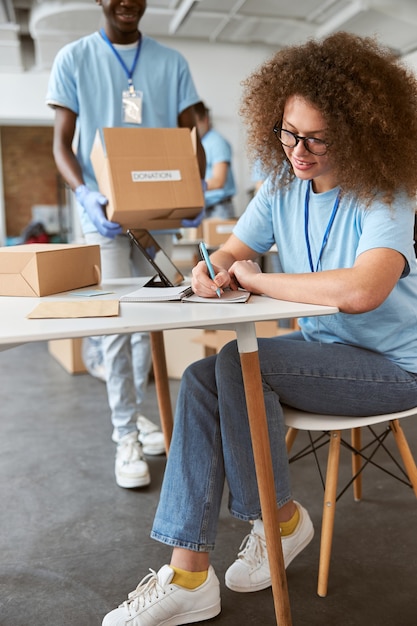 woman making notes and guy carrying cardboard box while working on donation project indoors