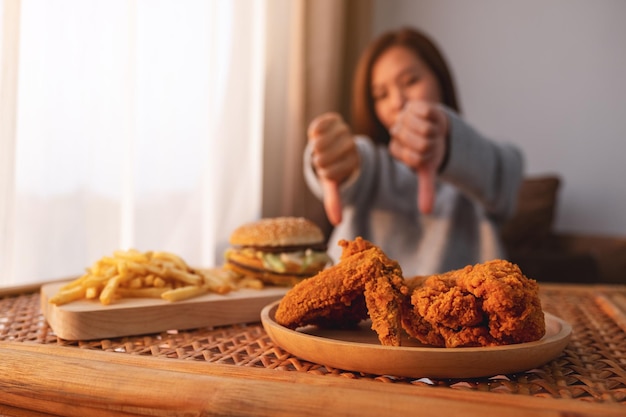 Женщина делает большой палец вниз знаком гамбургеру, картофелю фри и жареному цыпленку на столе для диеты и концепции здорового питания