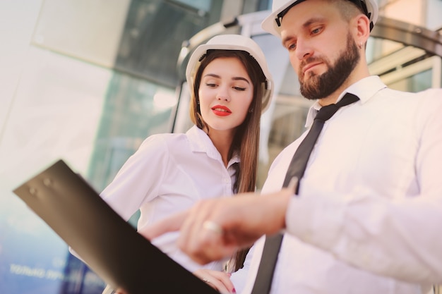 A woman and a man in business clothes and in white construction helmets discuss a construction plan 