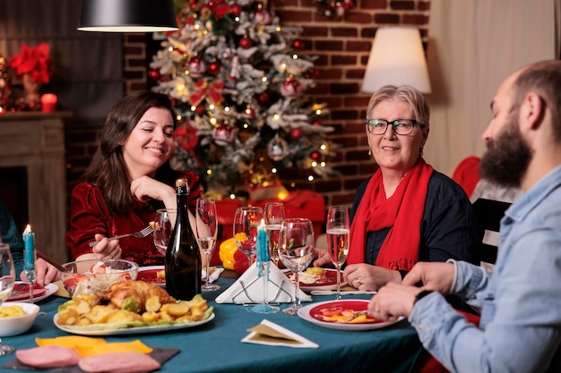 Женщина и мужчина празднуют рождество с родителями, семья сидит за праздничным обеденным столом на домашней вечеринке. Люди едят традиционную рождественскую еду, пьют игристое вино, смеются