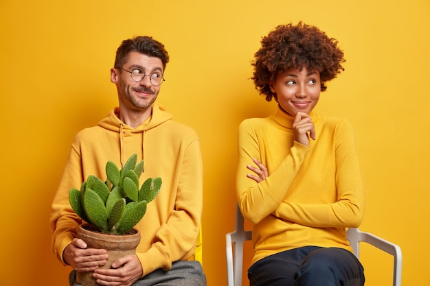 woman and man sit closely to each other on comfortable chairs guy holds pot of cactus isolated on yellow