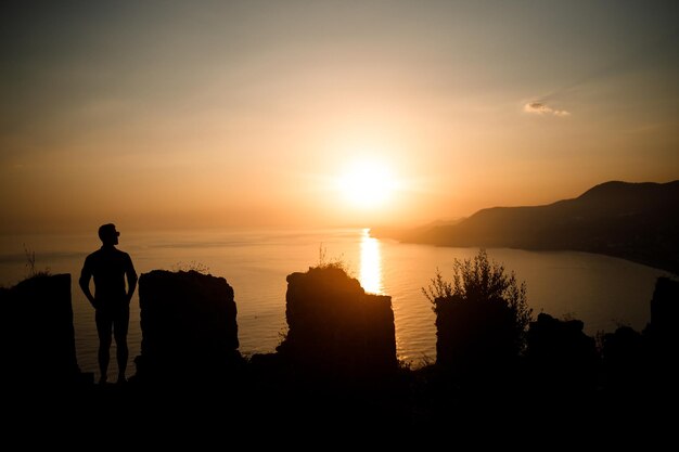 Женщина или мужчина, стоящие на скале и смотрящие прямо на закат у моря Концепция природы и красоты Оранжевый закат Силуэт на закате