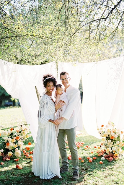 Woman next to a man with a baby in his arms near a white curtain in the garden