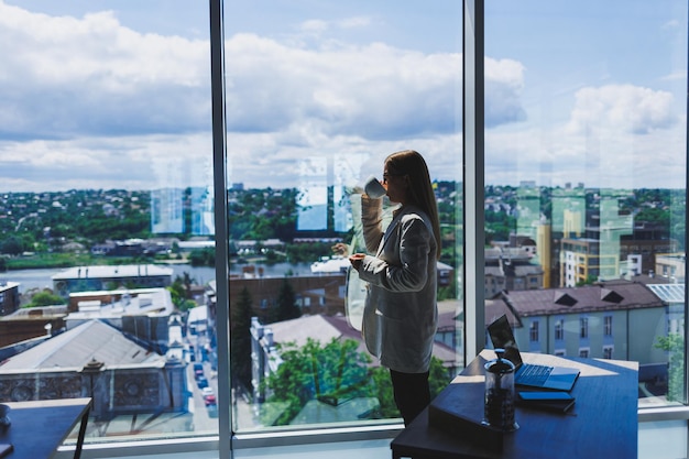 Woman manager in a jacket and glasses on her face drinking coffee during a break at work standing with a cup in the office pensive female project planning freelancer doing remote work