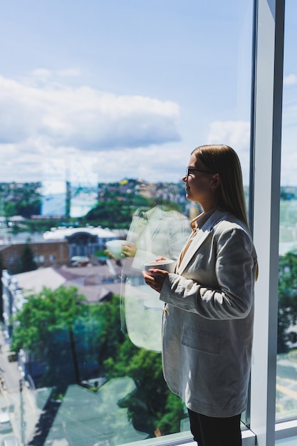 Woman manager in a jacket and glasses on her face drinking coffee during a break at work standing with a cup in the office pensive female project planning freelancer doing remote work