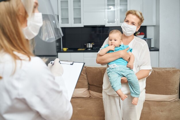 Woman in mask carrying boy while doctor writing on clipboard