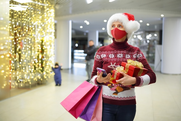 クリスマス前のCovid19で買い物をしている間、マスクをした女性がギフトや買い物袋を持っています