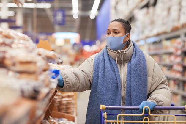 Woman in mask walking with shopping cart along shelves in grocery shop