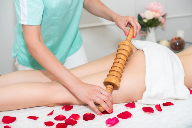 Женщина-массажист делает антицеллюлитный массаж молодой девушке, деревянный роликовый массажер. ноги накрыть белым полотенцем.