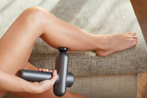 Photo a woman massages her legs with an automatic pistol at home. shock self-massage to restore fascia muscles and trigger points