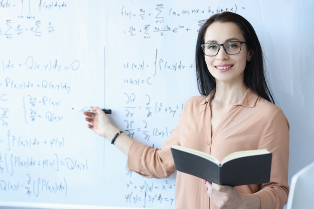 Matematico donna con gli occhiali in piedi alla lavagna con formule e tenendo libro aperto