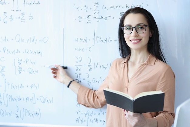 Woman mathematician with glasses standing at blackboard with formulas and holding open book