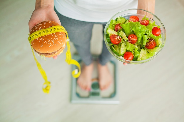 Foto peso corporeo di misurazione della donna sull'hamburger e sull'insalata della tenuta della bilancia.