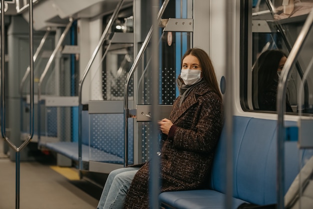 A woman in a medical face mask to avoid the spread of coronavirus is sitting staring to the side in a modern subway car. A girl in a surgical mask against COVID-19 is taking a ride on a metro train.