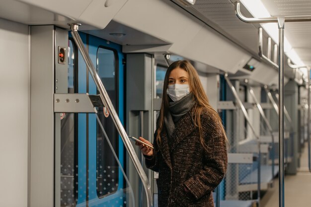 A woman in a medical face mask to avoid the spread of coronavirus is standing near doors and staring to the side in an empty subway car. A girl in a surgical mask is taking a ride on a metro train.