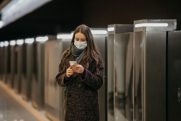 코로나바이러스 확산을 피하기 위해 의료용 안면 마스크를 쓴 여성이 지하철 승강장에서 스마트폰을 사용하고 있습니다. 수술용 마스크를 쓴 소녀가 지하철에서 사회적 거리를 유지하고 있습니다.