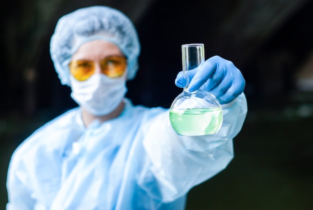 Woman in a medical or laboratory suit shows a flask with greenish liquid