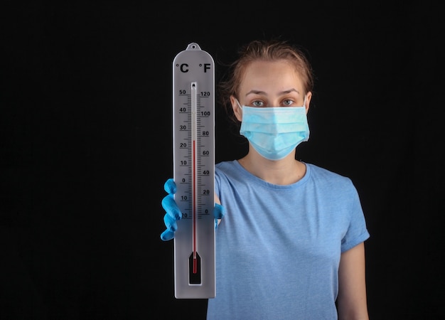 A woman in a medical mask and gloves holds a weather thermometer