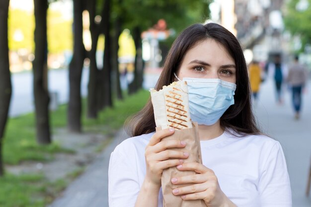 A woman in a medical mask keeps shawarma on the street.