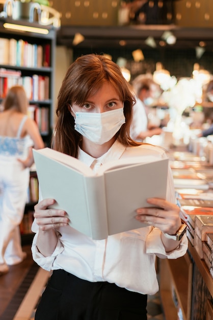 Женщина в медицинской маске в библиотеке, держа в руках книгу