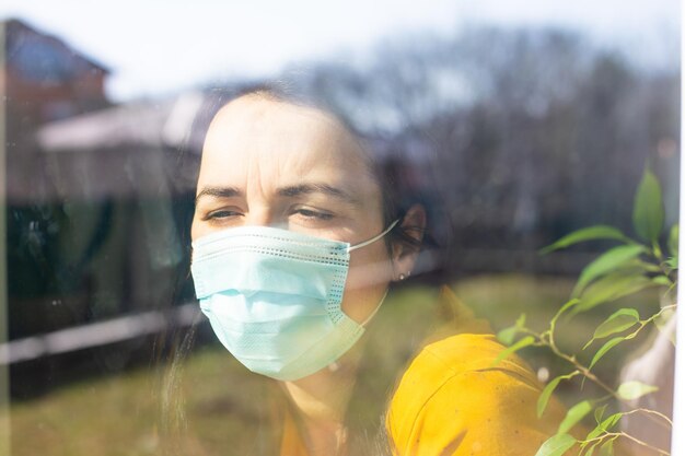 Женщина в медицинской маске смотрит в окно
