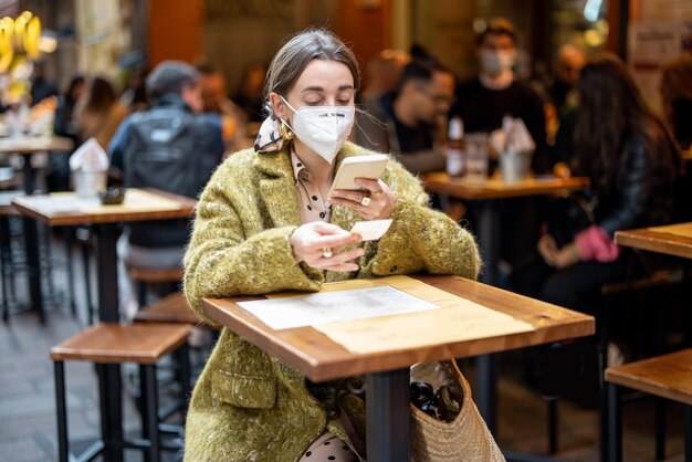 Woman in medical mask photographing qr code of restaurant menu outdoors