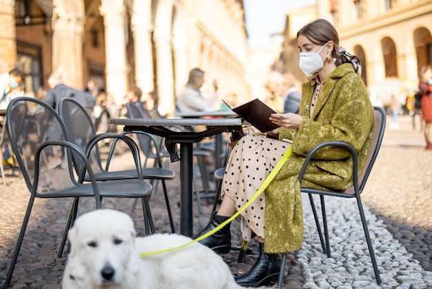 古いイタリアの都市の屋外カフェで彼女の犬と一緒に座っている医療マスクの女性