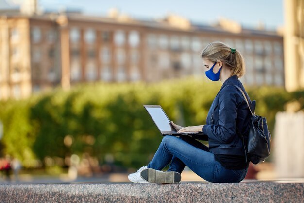 Woman in medical mask with laptop sits outdoors at daytime