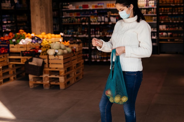 Woman in medical mask with mesh bag full of fruits checking the receipt while shopping Personal accaunting to controll expanses Rational money spending and concious buying while pandemic