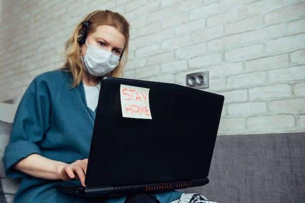 自宅からオンラインのラップトップに取り組んで医療マスクの女性。ウイルスの大流行の間、家にいること。検疫、自己分離、疾病予防のコンセプト。テキスト付きステッカー