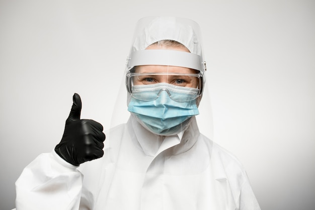 Woman in medical protective clothing and black latex glove shows hand thumbs up sign.