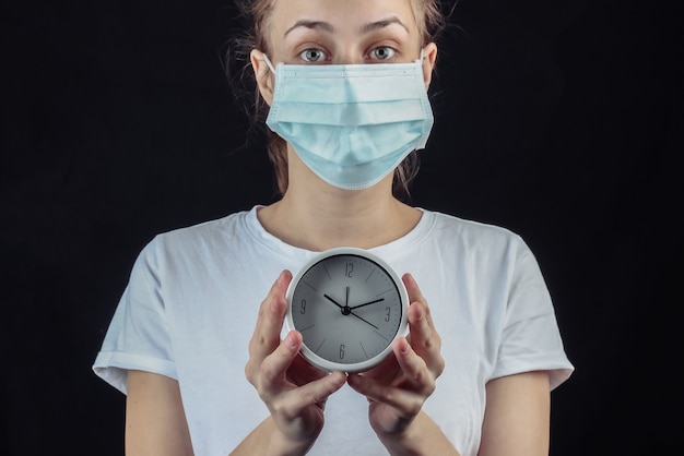 Woman in a medical protective mask holding white clock on a black wall.