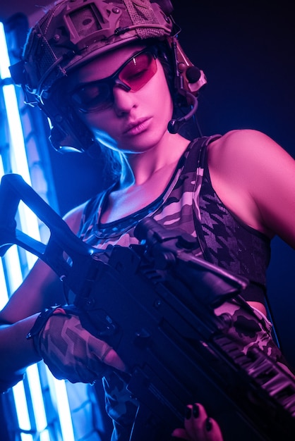 Donna in tuta militare softair in posa con una pistola in mano su un muro scuro nella foschia nella luce al neon