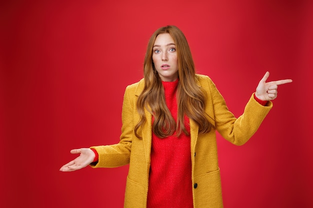 Woman missed client posing confused and unaware how happened shrugging making clueless gesture with palm as pointing right and looking questioned and uncertain at camera over red background.