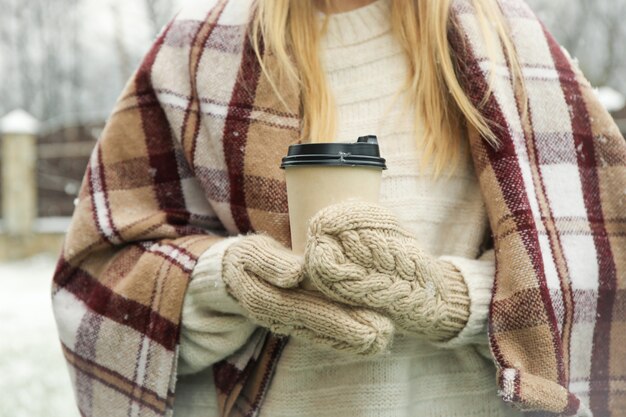 雪の降る天気でコーヒーを保持しているミトンと格子縞の女性