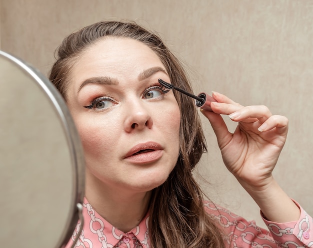 A woman in the morning at home paints her eyes while looking in the mirror.