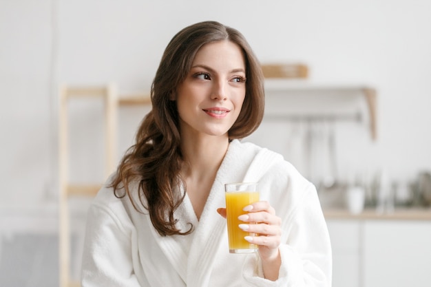 手に新鮮なオレンジジュースと朝の女性。朝食にオレンジジュースを飲む白いバスローブの女性ポジティブ完璧な朝の女性の肖像画。スタジオショット。