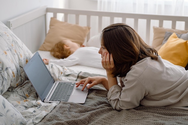 Woman mother using laptop lying on the bed while child sleeps in the crib next to her Working mom