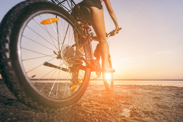 The woman in a multicolored suit sits on a bicycle in a desert area near the water. Fitness concept. Rear view and bottom view. Close-up