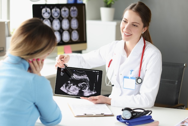 Woman obstetrician gynecologist showing patient photograph of ultrasound examination of fetus on digital tablet