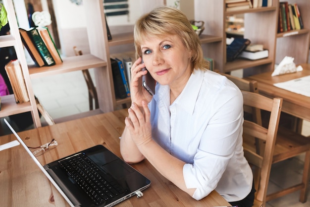 A woman in the office is working on a laptop. She's on the phone. A middle-aged woman, an adult in a cafe.